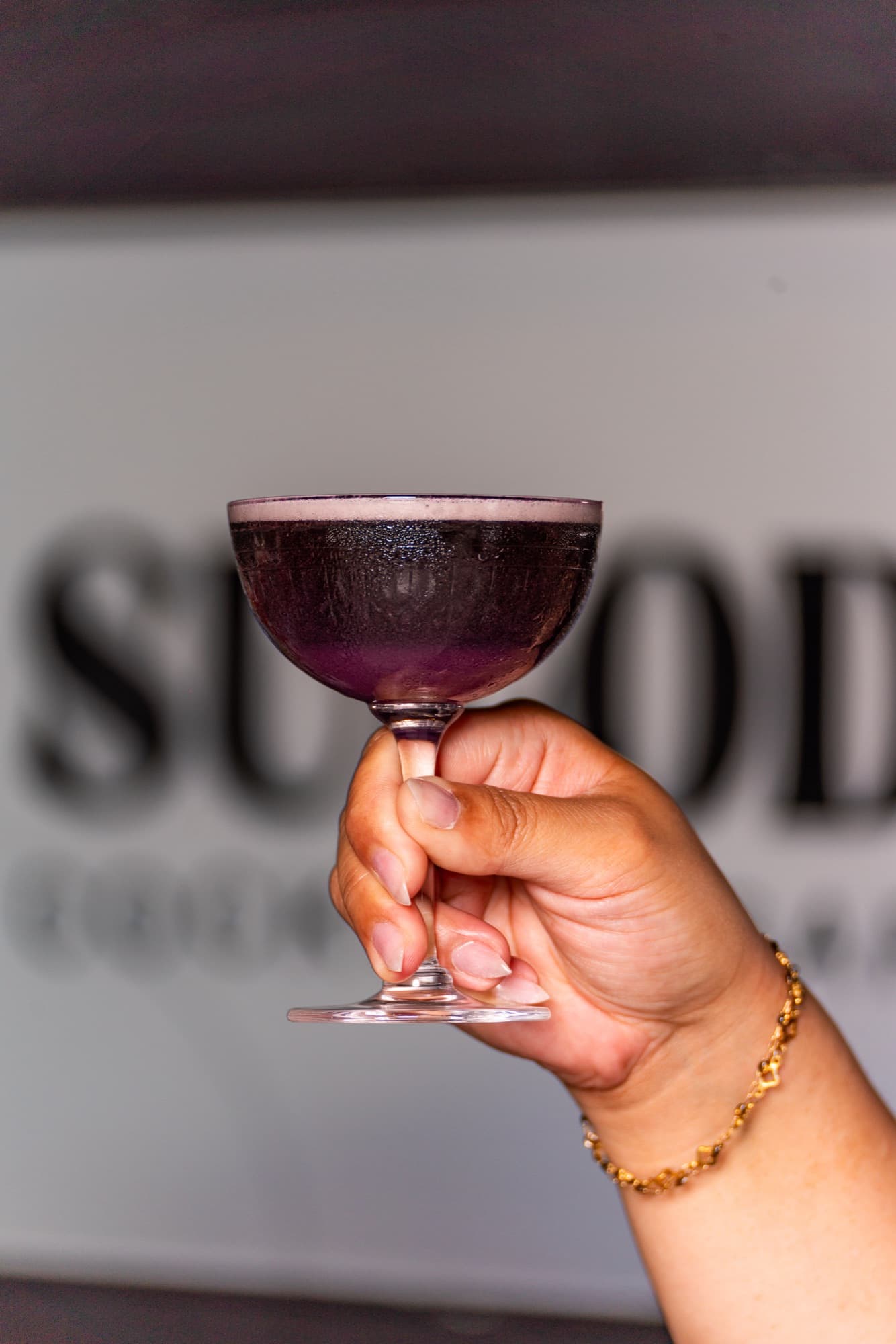 The Purple Pick Me Up, a deep purple ube cocktail in a coupe, raised in front of the Sulod sign