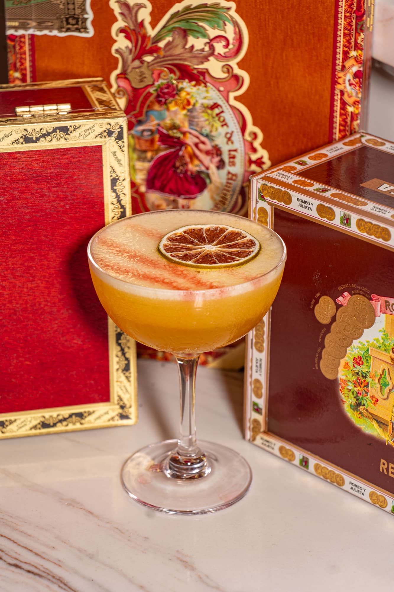 A golden coupe with a dried lime wheel, staged among ornate cigar boxes