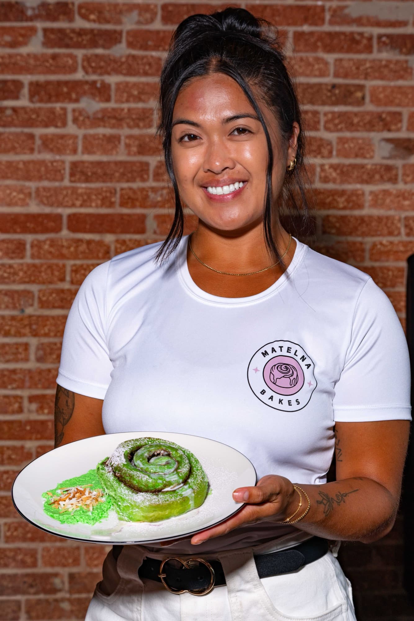 Matelna in her bakery whites, holding a pandan cinnamon roll dusted with sugar